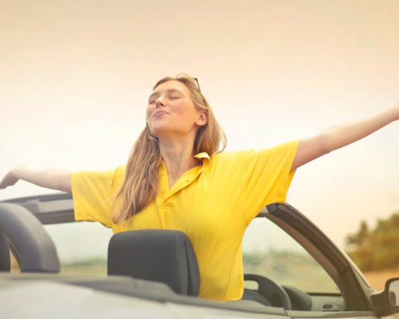 A cheerful woman in a yellow shirt enjoys freedom on a sunny drive in a convertible.